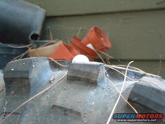 Close-up of a tire tread with pine needles and a white ball on top. Some orange plant pots are in the background