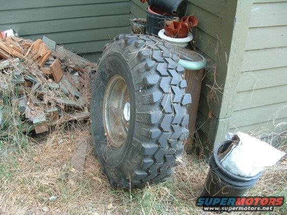 Off-road tire with aggressive tread leaning against a weathered green building. Pile of wood scraps to the left