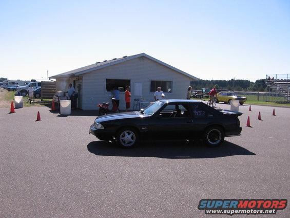 Black 1989 Ford Mustang coupe parked outside a building at a track, with racing numbers on the door