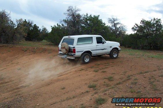 White Ford Bronco driving on a dirt road, kicking up dust. Spare tire mounted on the back