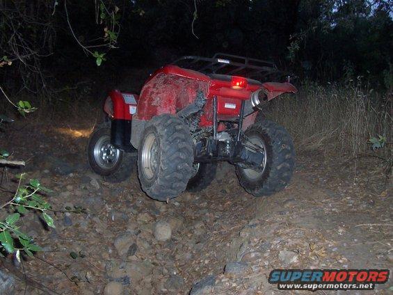 Red ATV on rocky terrain, rear view, with large knobby tires and a cargo rack