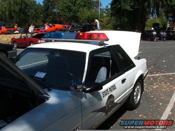 White Ford Mustang police car with light bar, parked at a car show with other vehicles