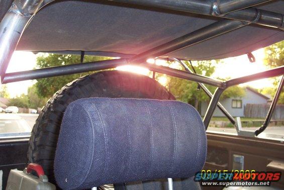 Interior view of a vehicle with a custom roll cage, spare tire, and headrest. Sunlight streams through the rear