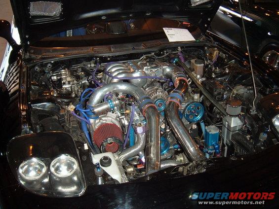Black car engine bay with twin turbochargers, chrome piping, and blue accents. A white paper is visible on top