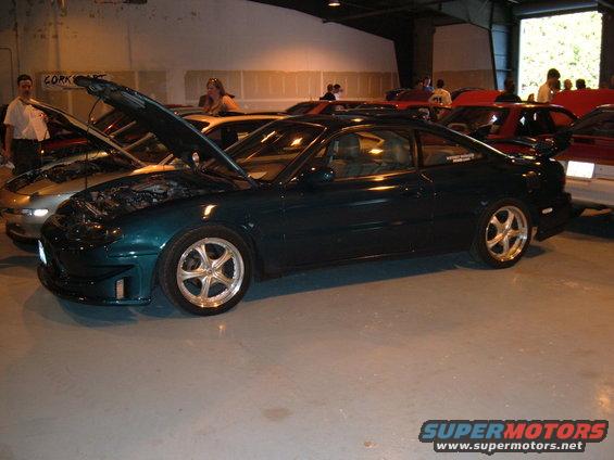 Dark green coupe with hood open, parked indoors next to other cars. Has aftermarket wheels