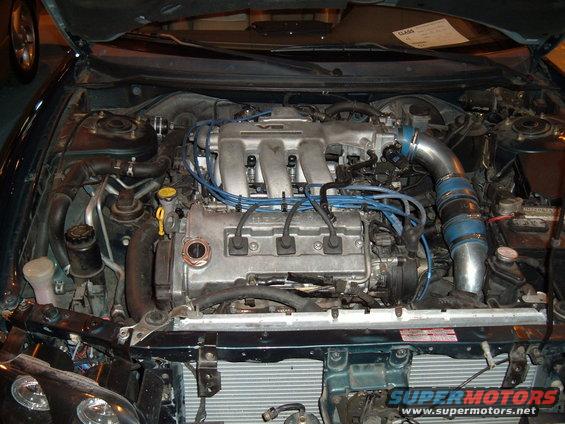 Engine bay of a 1991 Ford Probe, V6 engine with blue spark plug wires, silver intake piping, and a white label on the hood