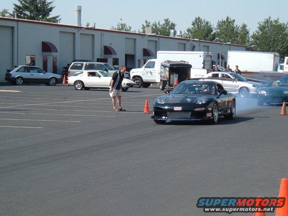 Black Mazda RX-7 FD doing a drift with smoke coming from tires. Other cars and buildings in background