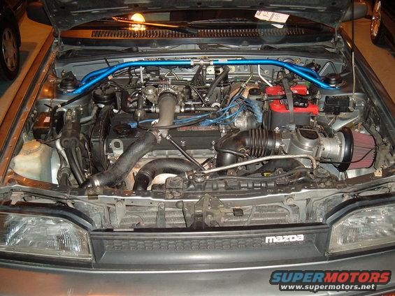 Engine bay with blue strut bar, red battery, and aftermarket air intake. Mazda logo visible on grille