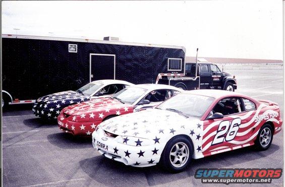 Three 1995 Ford Mustangs with patriotic star and stripe paint jobs parked in front of a trailer and a flatbed truck