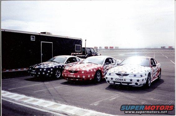 Three 1995 Ford Mustangs with star and stripe paint jobs parked on asphalt. The middle car has a red base with white stars, the left car has a dark base with white stars, and the right car has a white base with red and blue stars and stripes. All have ...