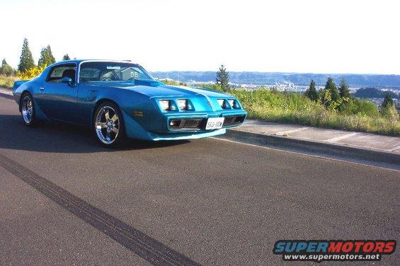 Blue 1977 Pontiac Firebird coupe with chrome wheels, hood scoop, and custom front end, parked on asphalt