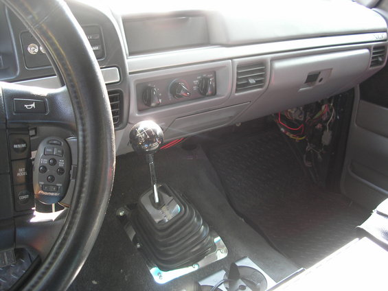 Vehicle interior showing dashboard, steering wheel with cruise control buttons, and a manual transmission shifter. Wires are exposed under the dashboard