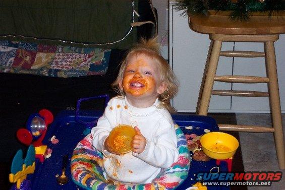 Toddler smeared with orange food sits in a blue baby walker with toys