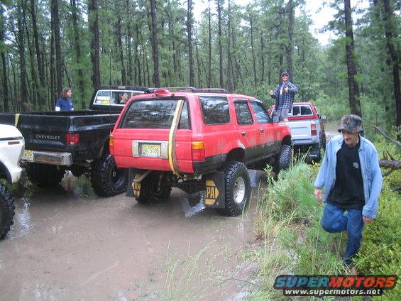 Red SUV stuck in mud, with a black Chevrolet pickup and a white pickup behind it. People are standing around the vehicles