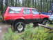 1990 Ford Bronco - A red SUV navigates through a muddy, wooded area with a pickup truck nearby.