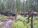 1990 Ford Bronco - Two military trucks, one green and one camouflage, are parked in a muddy forest with trees all around.