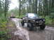 1990 Ford Bronco - Two trucks navigate a wet, muddy path through a dense forest.