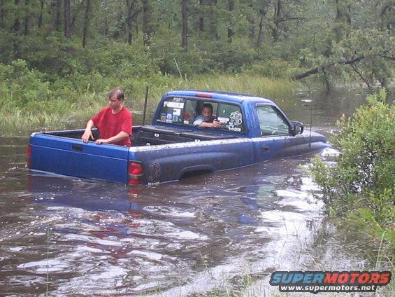 Blue pickup truck submerged in water with two people inside and one in the bed