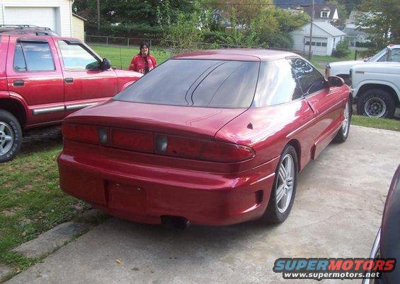 Red coupe with tinted windows, rear view, parked on concrete driveway next to a red SUV and white truck