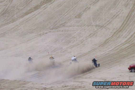 Four three-wheeled vehicles kick up dust as they ride down sand dunes. A red SUV is parked on the right