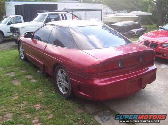 Red coupe parked in a yard with a white truck and boats in background. Supermotors.net logo visible