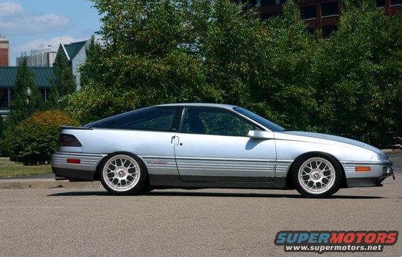 Silver coupe with aftermarket wheels parked on pavement, trees and buildings in background