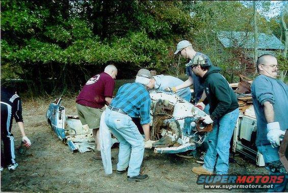 Several men are gathered around the front end of a heavily dismantled vehicle, possibly working on its removal or salvage