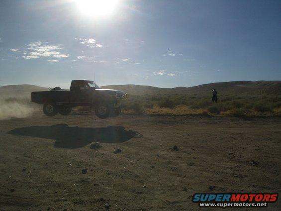 Truck airborne over dirt road, kicking up dust. Hills and a person in the background