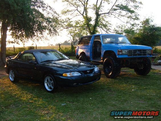 Blue lifted 1987 Ford Bronco with driver's door open, parked next to a dark green convertible Mustang