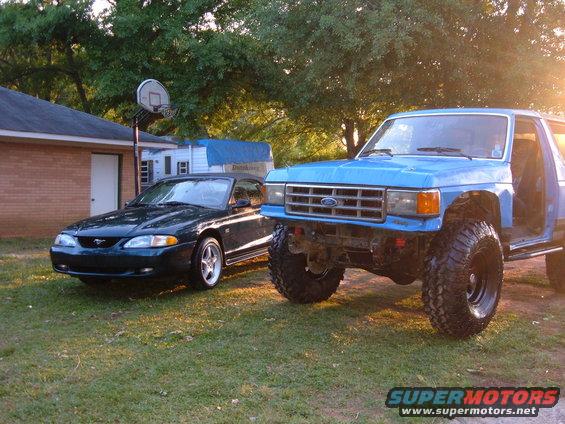 Blue lifted Ford truck with large tires parked next to a dark green Ford Mustang in a grassy yard
