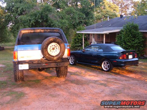 Rear view of a blue and white Ford Bronco with a spare tire on the back, parked next to a dark green Ford Mustang convertible