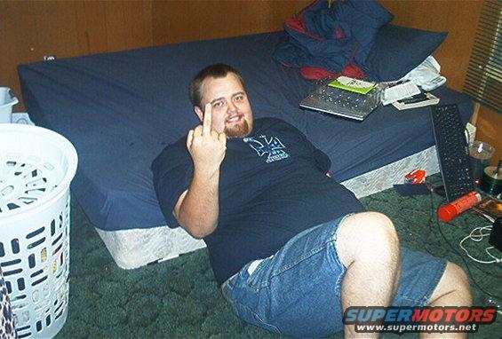 Man in a t-shirt and denim shorts lies on a bed, giving the middle finger