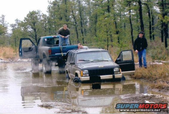 1997 Jeep Cherokee stuck in mud, front end submerged. Blue pickup truck with open door behind it