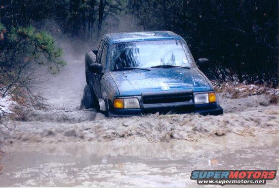 Blue pickup truck driving through a muddy water crossing with trees in the background