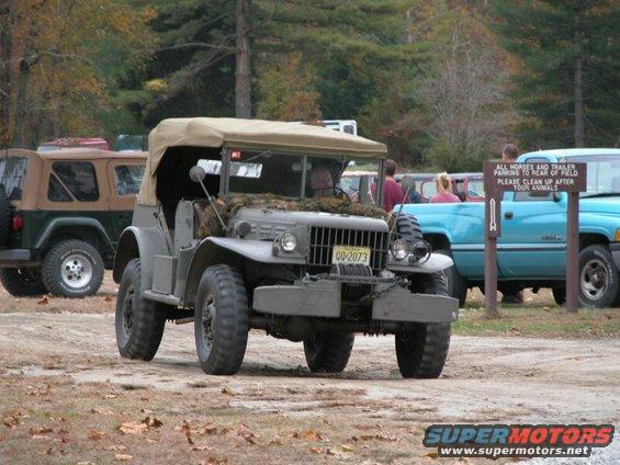 Olive drab military truck with a tan canvas top, front view, parked on a dirt lot with other vehicles