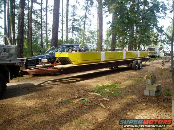 Tow truck pulling a trailer loaded with long yellow cargo; RV in background