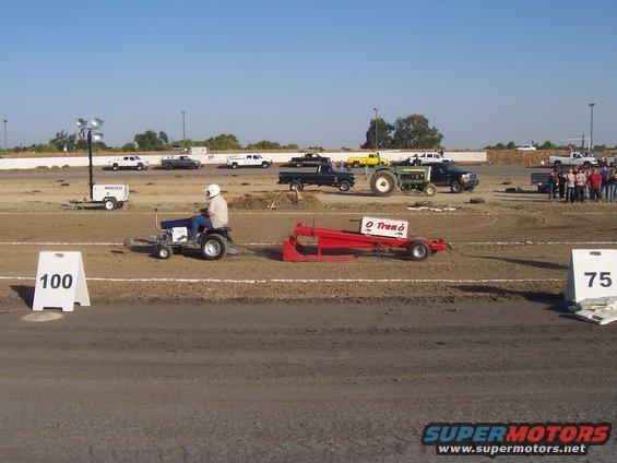 Lawn tractor with driver pulls red sled on dirt track; white signs show 100 and 75