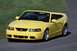 2003 Ford Mustang - A yellow 2003 Ford Mustang convertible is captured in motion on a paved road with a grassy embankment in the background.