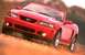 2003 Ford Mustang - A red 2003 Ford Mustang convertible is captured in a dynamic shot as it drives on a dirt road, kicking up dust.