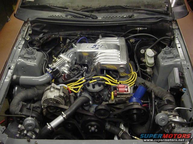 Engine bay with a V8 engine, chrome intake manifold, and yellow spark plug wires. Supermotors.net logo visible