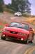 2003 Ford Mustang - A red 2003 Ford Mustang convertible cruises down a scenic, winding road with another vehicle in the distance.