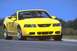 2003 Ford Mustang - A bright yellow 2003 Ford Mustang convertible is captured in a three-quarter front view, driving on a paved road under a clear blue sky.
