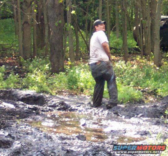 Man in dirty jeans and white t-shirt stands in a muddy, wooded area with puddles