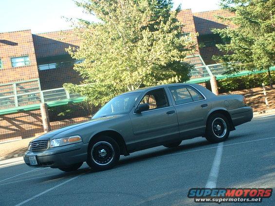 2000 Ford Crown Victoria, gray, parked on asphalt with a brick building and trees in background