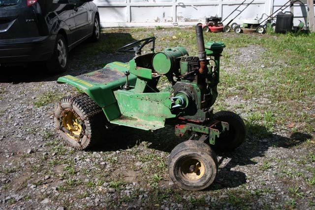 Green John Deere lawn tractor with chains on rear tire, parked on gravel next to a black car
