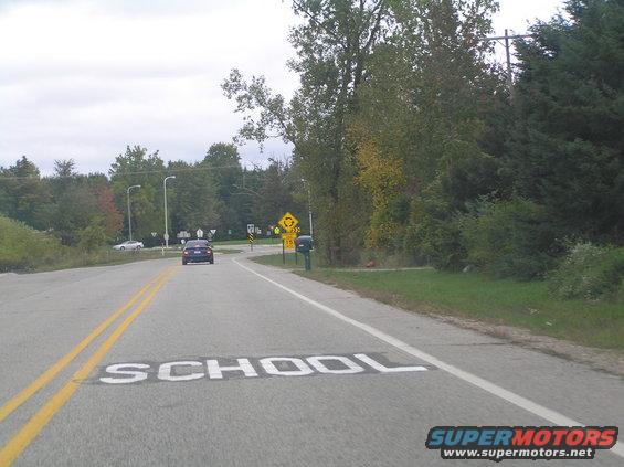 Car driving on road with 'SCHOOL' painted on it, approaching a roundabout