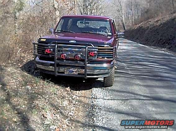Red Ford Bronco with a large chrome grille guard parked on a dirt shoulder next to a paved road