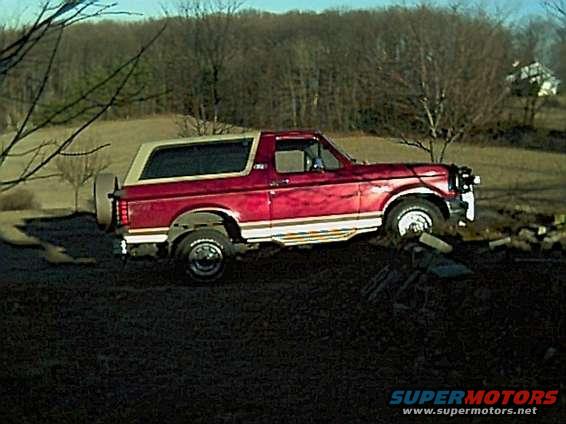 Red Ford Bronco parked on uneven ground, trees and a house in background