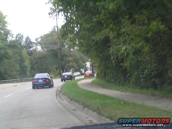 Blue sedan from rear, driving on a road with trees and a sidewalk