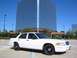 1998 Ford Crown Victoria - A white 1998 Ford Crown Victoria sedan is parked in a lot with a modern office building in the background.